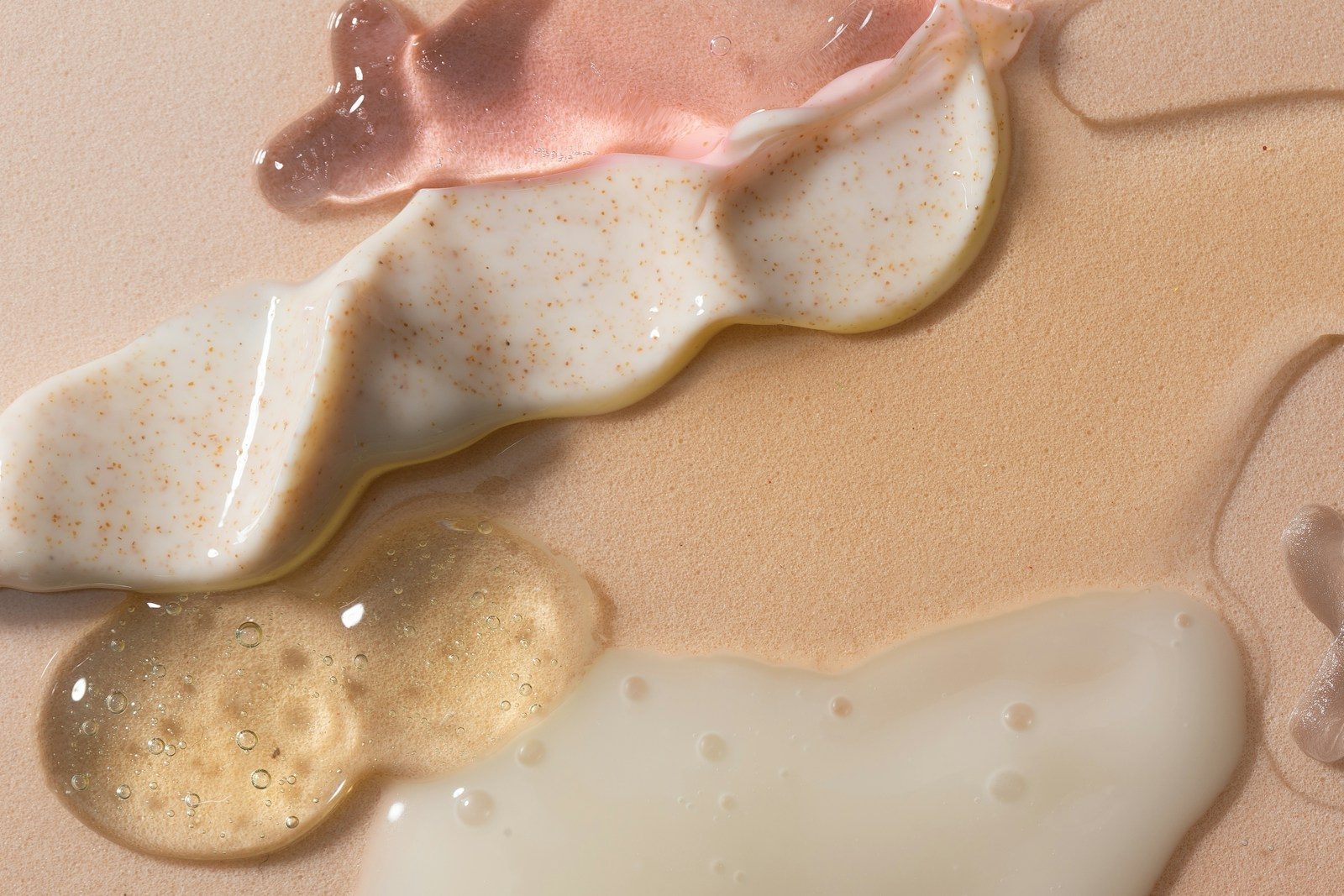 Swirls of pink gel, white exfoliating cream, and clear serum on a beige surface
